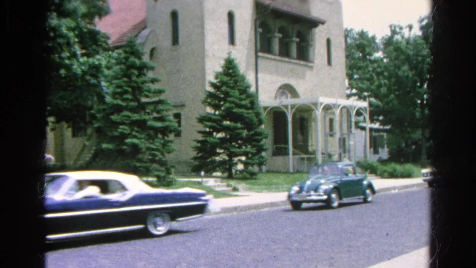 1966 Danville, Illinois home-movie frame with a dark convertible and blue car passing the church exterior on a sunny street.