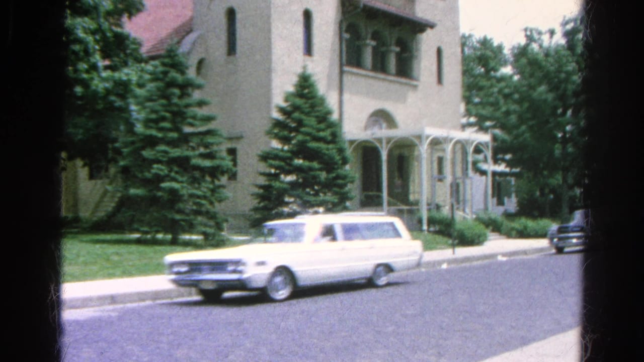 White station wagon passes a large building with arched windows and evergreen trees.