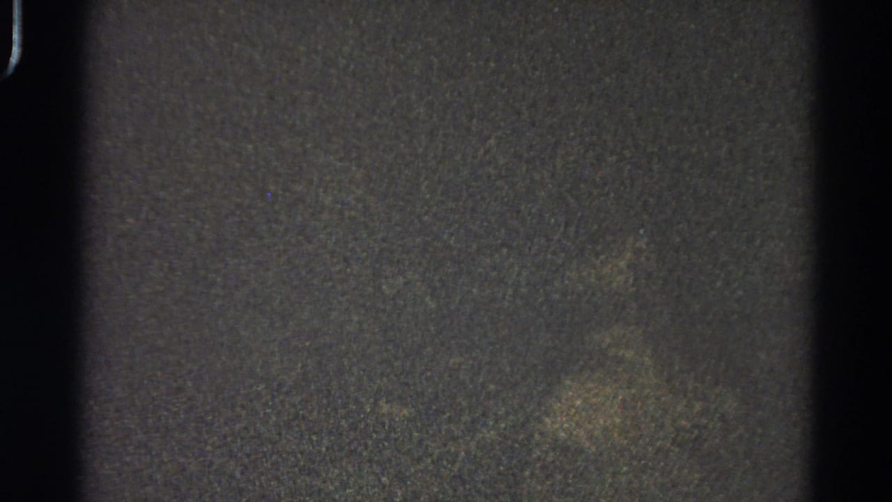 Very dark grainy frame with black film edge and faint mottled shapes.