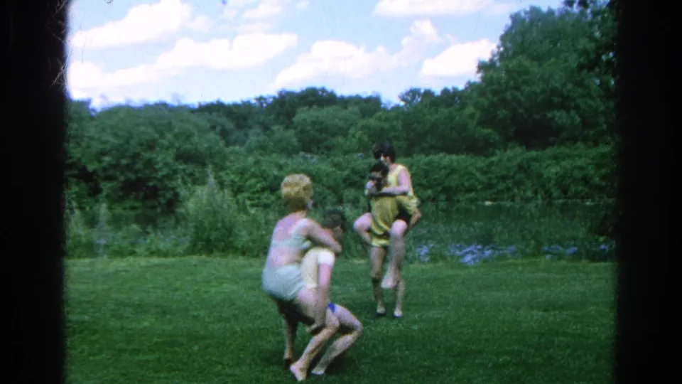 Two pairs continue playful lifting or carrying on a lawn near a wooded pond in 1966 Danville, Illinois footage.