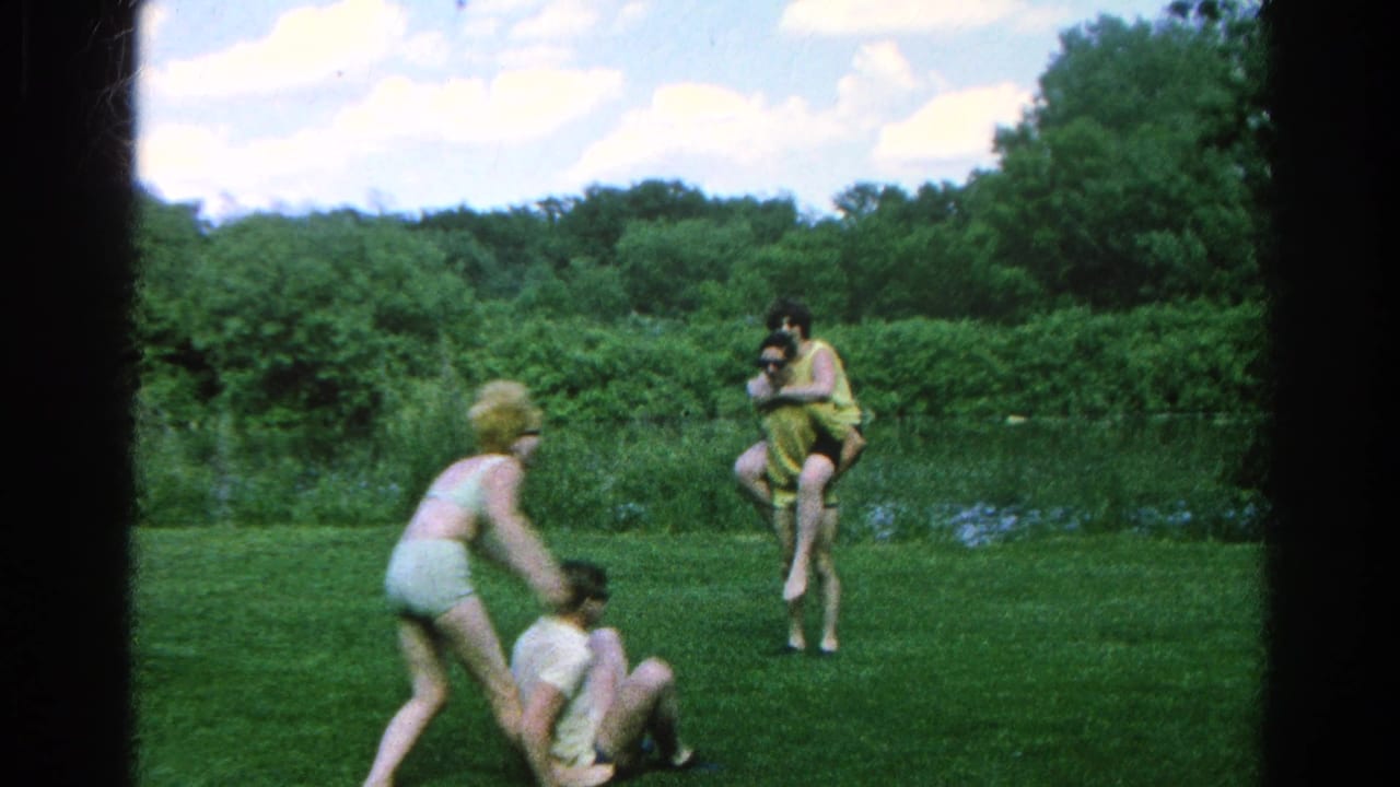 Young adults play on a green lawn near a pond and treeline.