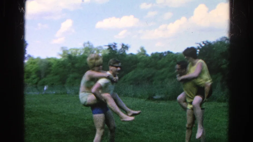 1966 home-movie frame from Danville showing two young-adult piggyback pairs facing each other on grass near trees and water.