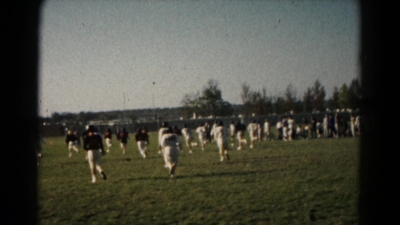Groups of players run across the field while spectators stand along the far sideline.