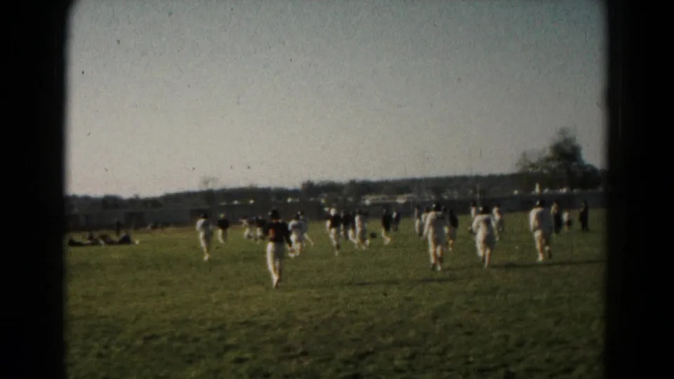 Wide home-movie frame of 1966 Hagerstown field play, with players moving across the grass and spectators at the edge.