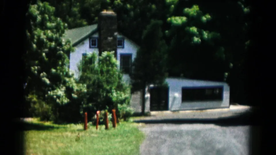 1966 Hagerstown-area home-movie frame showing a white house behind grass, trees, a driveway, garage, stone chimney, and red posts.