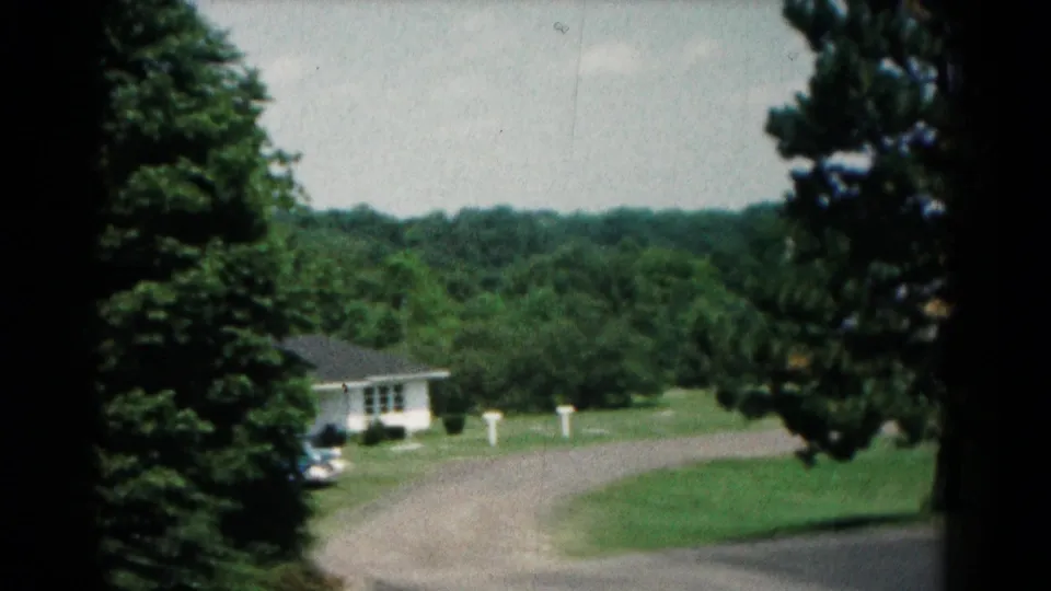 1966 Hagerstown home-movie frame showing a white house near a curved drive, parked car, lawn, and wooded backdrop.