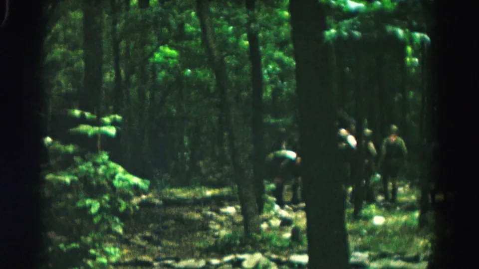 Darker 1966 8mm frame from the Hagerstown-area woods showing several people gathered and bending near trees and scattered gear.