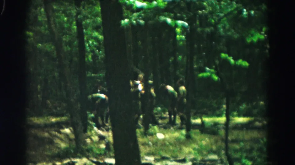 Soft 8mm frame showing a small group gathered in a shaded Maryland forest, with tree trunks and foliage obscuring details.