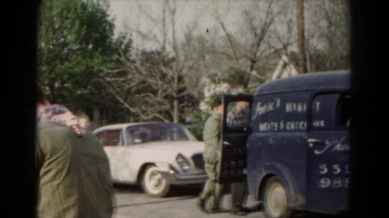 1966 Hagerstown home-movie frame showing a blue Frank's Market van, a white classic car, and adults near open vehicle doors.