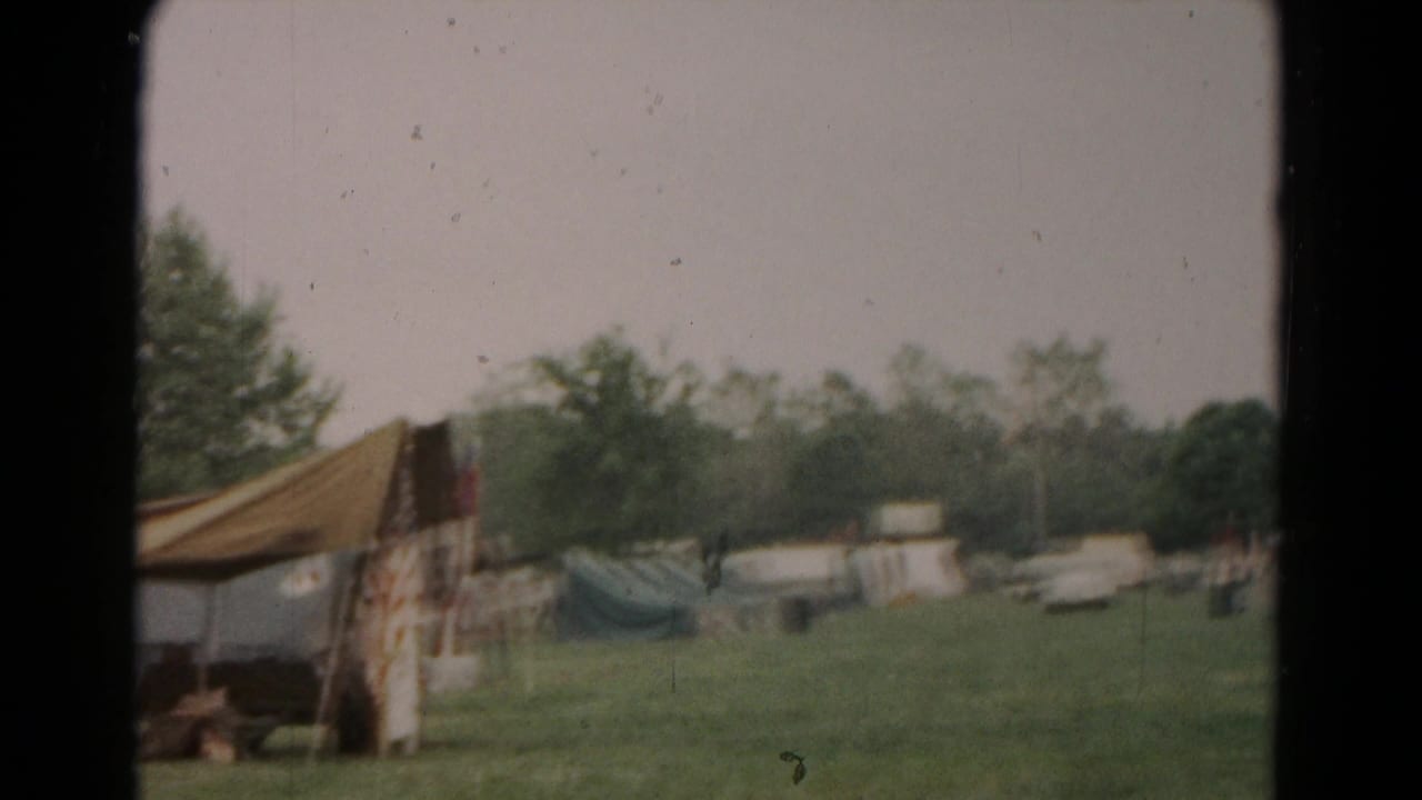 Grassy campsite with tents, trees, and a canvas awning at the edge of the frame.