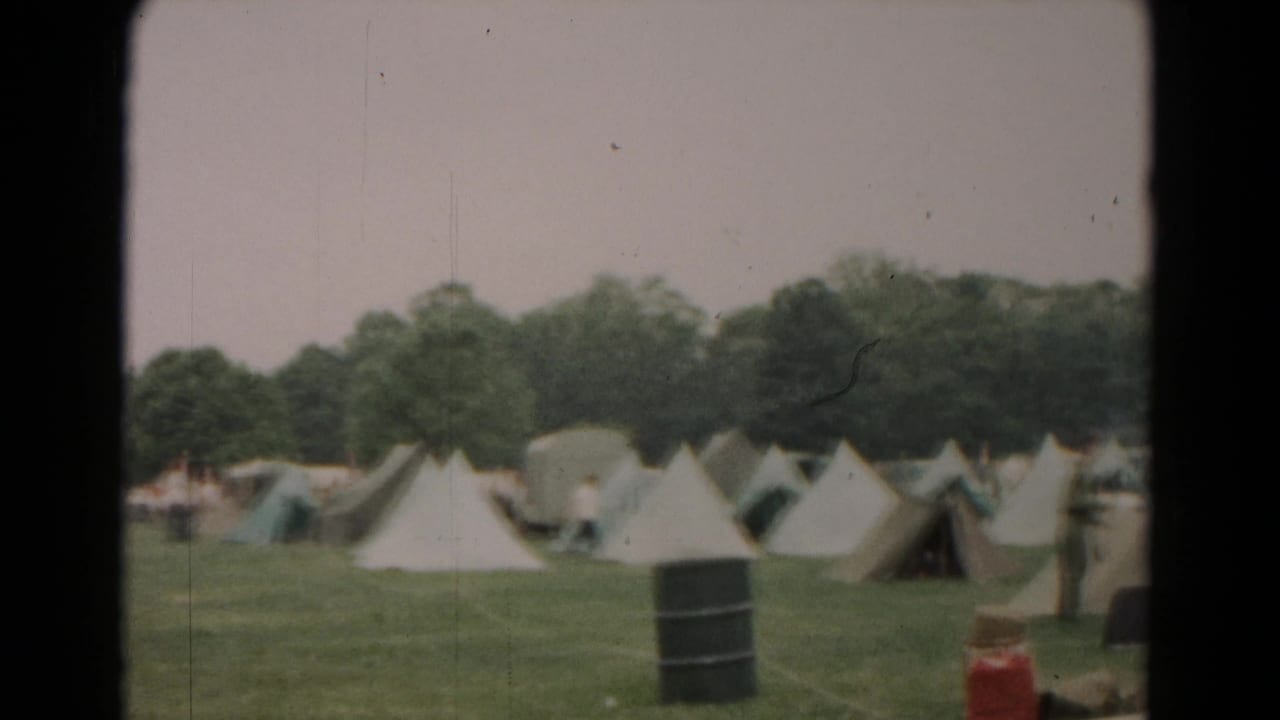 1966 Hagerstown campground frame showing rows of tents, trees, a dark barrel, and camping gear in the foreground.