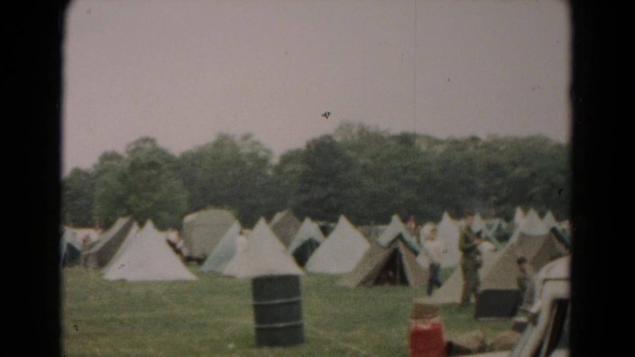 1966 Hagerstown home-movie frame showing canvas tents in rows, men in the field, a black barrel, and trees beyond the camp.