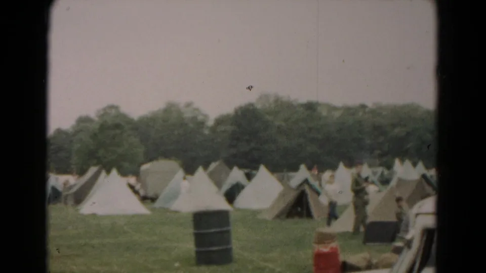1966 Hagerstown home-movie frame showing canvas tents in rows, men in the field, a black barrel, and trees beyond the camp.