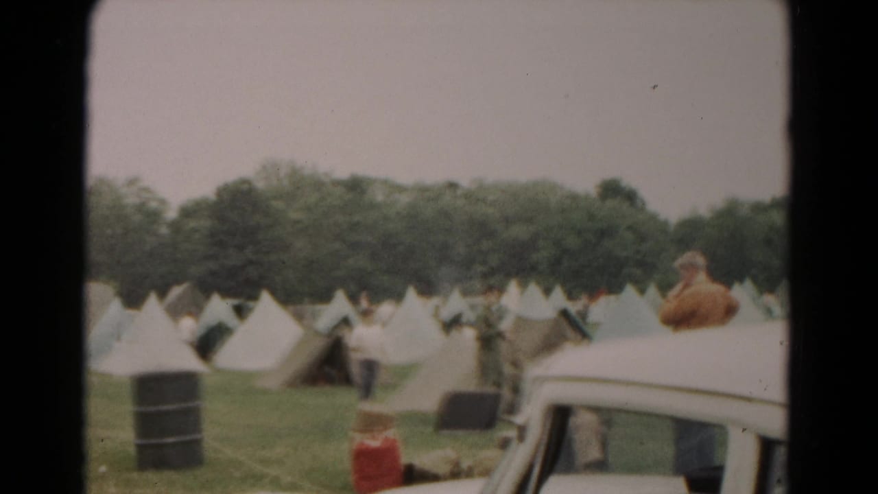 Tent rows and men in a field, with a white car partly visible at the right edge.