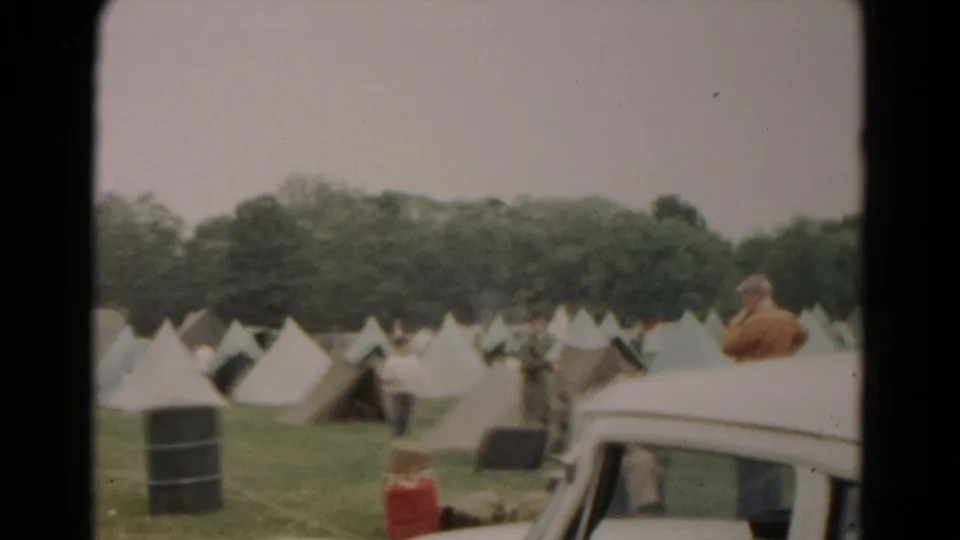1966 Hagerstown frame with pale tents, men walking through the encampment, a barrel, red container, and part of a white car.