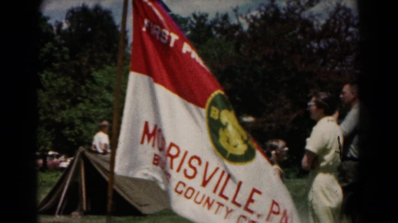 Red-and-white church flag beside canvas tents and adults on a grassy field.
