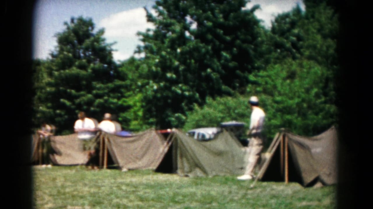 Hagerstown campsite view showing adults near canvas tents, with trees and parked cars behind the camp area.