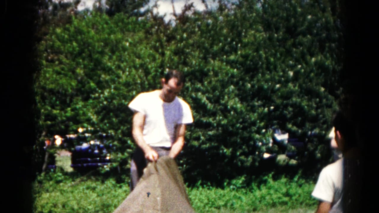 Man in a white T-shirt gathers a large brown sack in a green backyard.