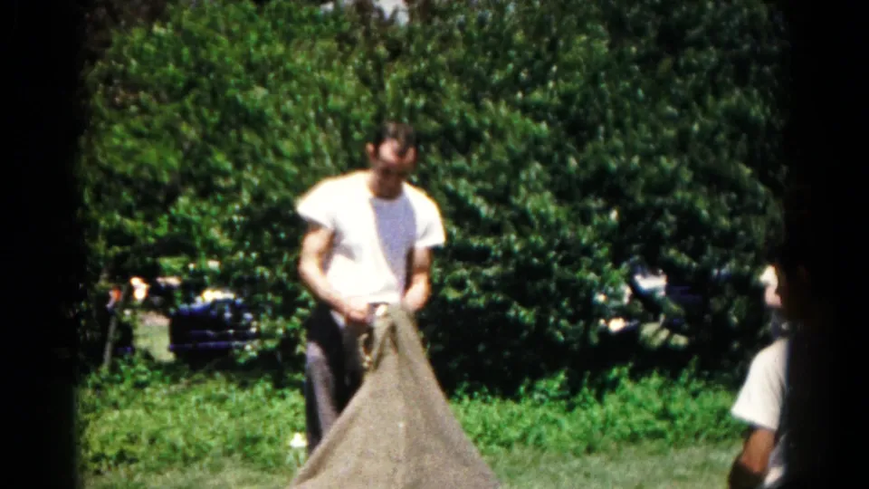 The man stands in the sun and works at the opening of a large sack, with shrubs and a partial parked car behind him.