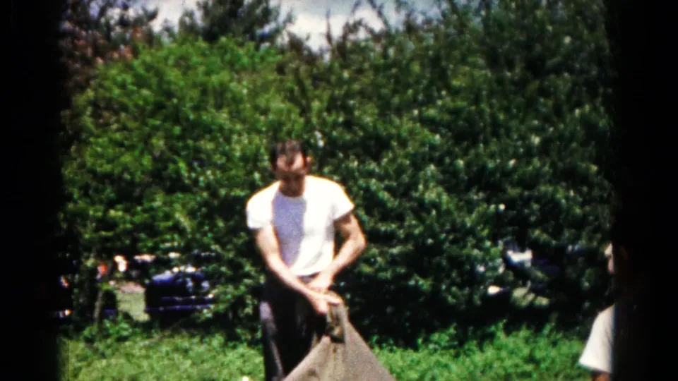 A man in a white T-shirt grips the top of a brown sack in a green Maryland backyard, likely during the 1966 home-movie scene.