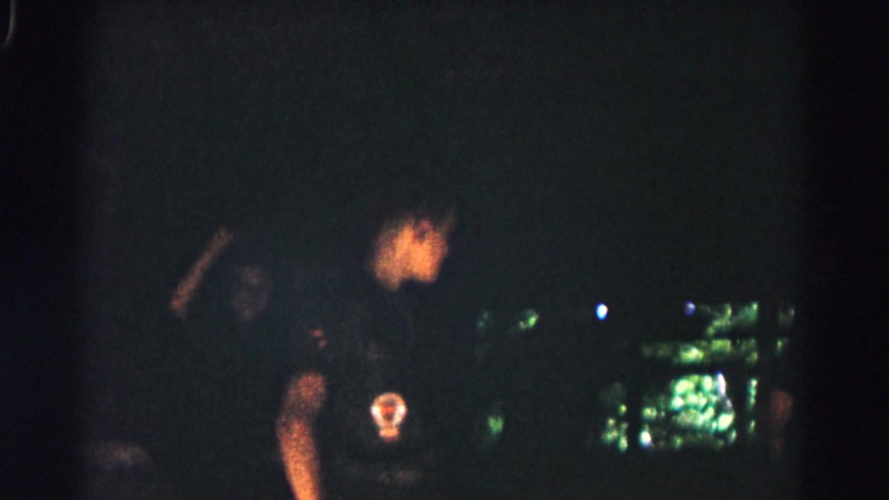 Dim nighttime frame with a person in dark clothing and a round chest patch near blurred lights.