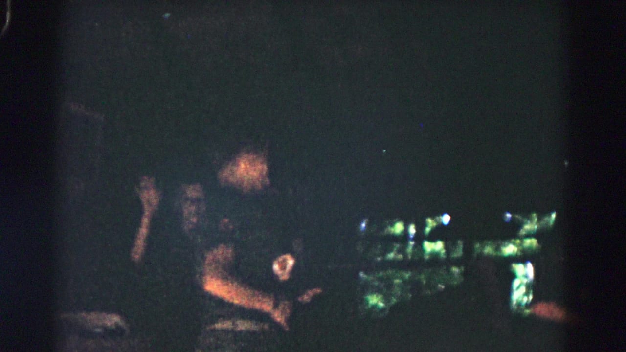 Person in dark clothing stands at night with a round chest patch and greenish lights behind them.