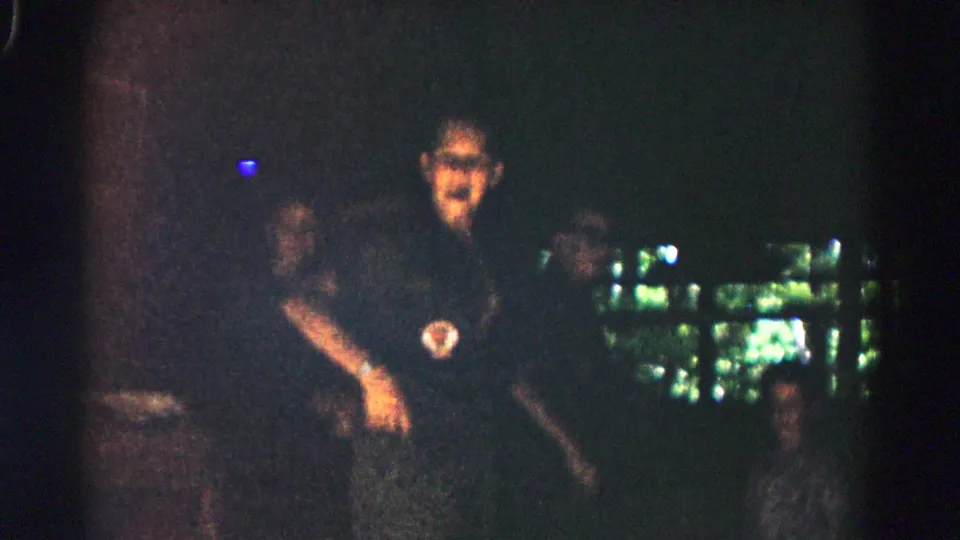 1966 Hagerstown footage shows a darkly dressed person with a round patch standing among shadowed people at a nighttime gathering.
