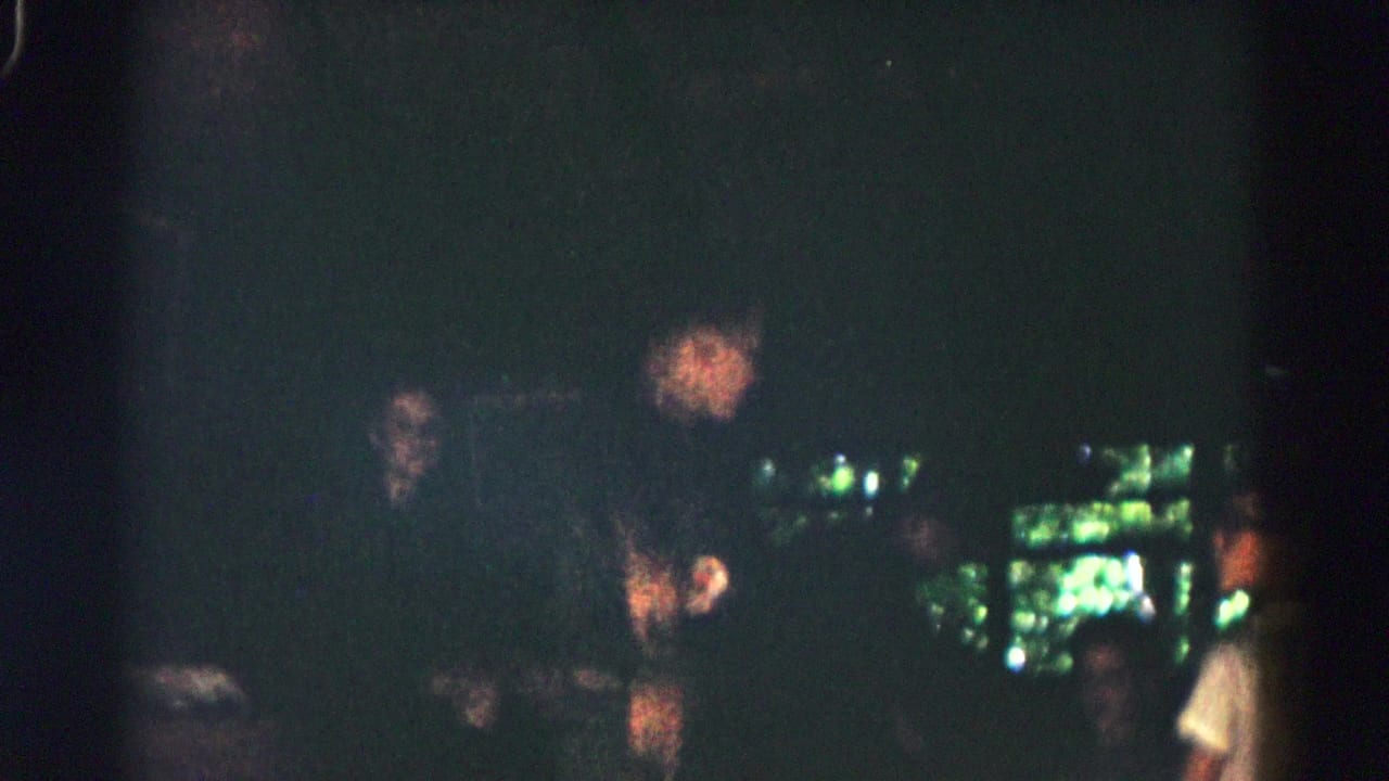 Dark interior or shaded scene with several indistinct people and bright green light in the background.