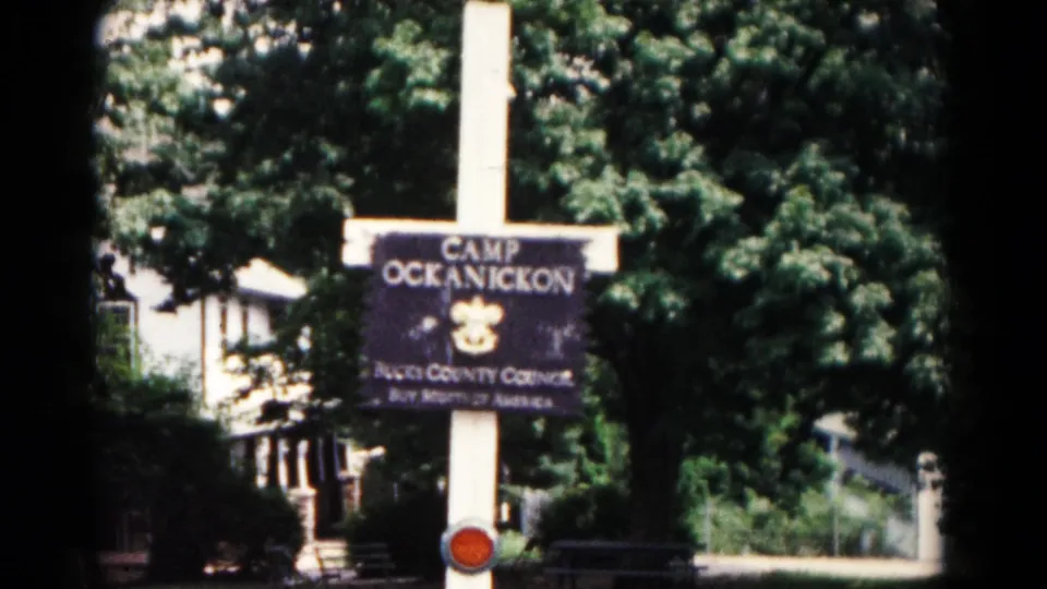 1966 Hagerstown frame with the Camp Ockanickon sign centered against dense trees, a pale structure, and a walkway.