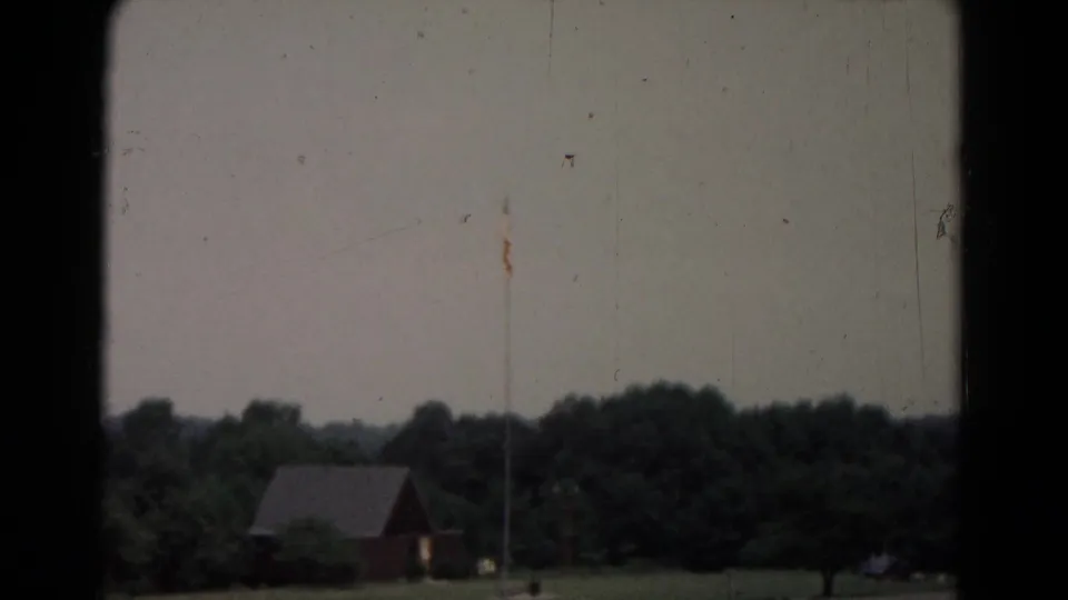 1960s 8mm home-movie frame showing a rural field, small red building, flagpole, and tree line through dark framing edges.