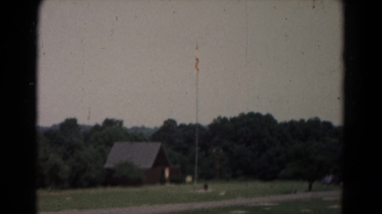 Archival 8mm frame of a rural landscape with a small red building, flagpole, open grass, and wooded background.