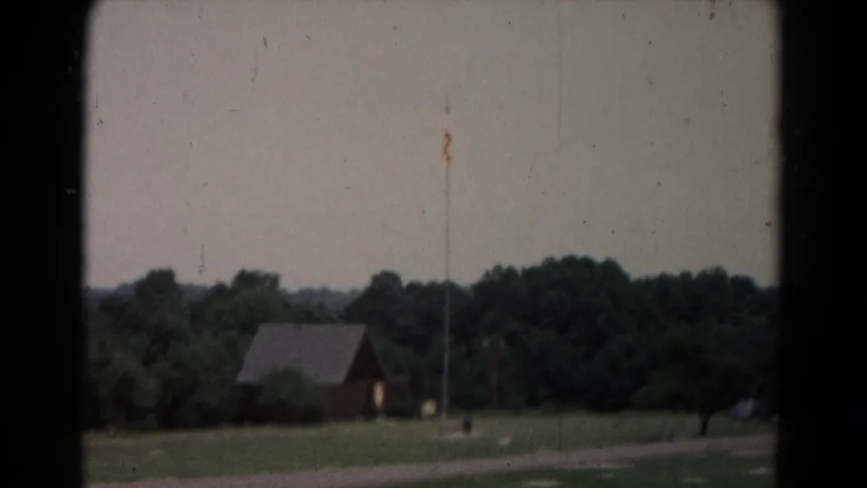Archival 8mm frame of a rural landscape with a small red building, flagpole, open grass, and wooded background.