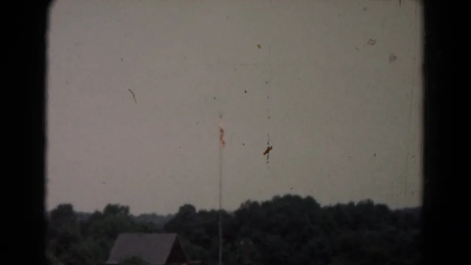 Soft 8mm view of a small rural building against trees, with visible dust and vertical film marks in the frame.