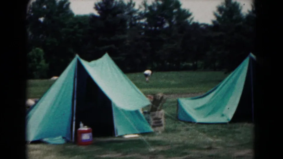 Green canvas tents stand in a wooded Hagerstown camp area, with a distant person visible in the field.