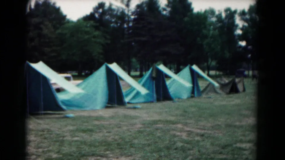 Wide 1966 Hagerstown, Maryland campsite frame with several green canvas tents stretching across grass before a wooded backdrop.