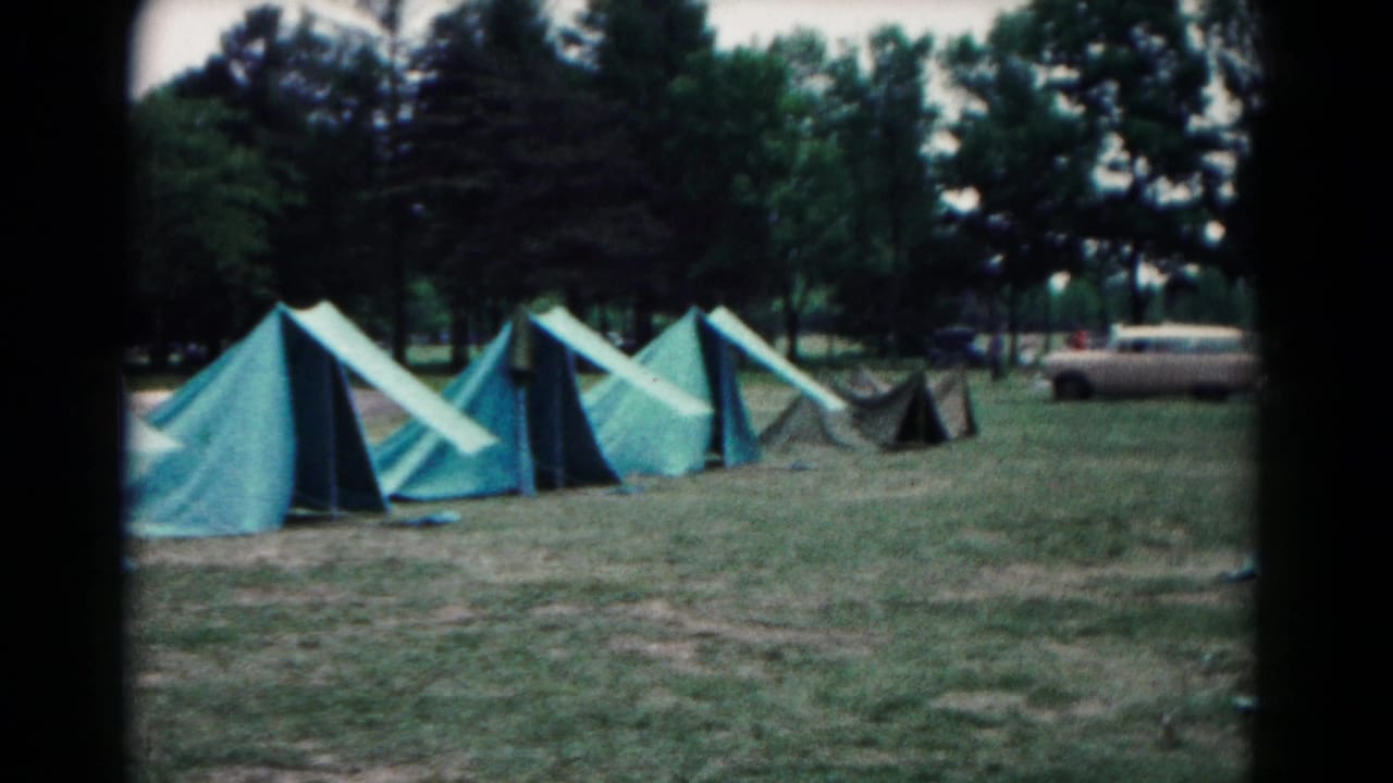 Rows of blue-green canvas tents on a grassy field with trees and a parked station wagon in the background.