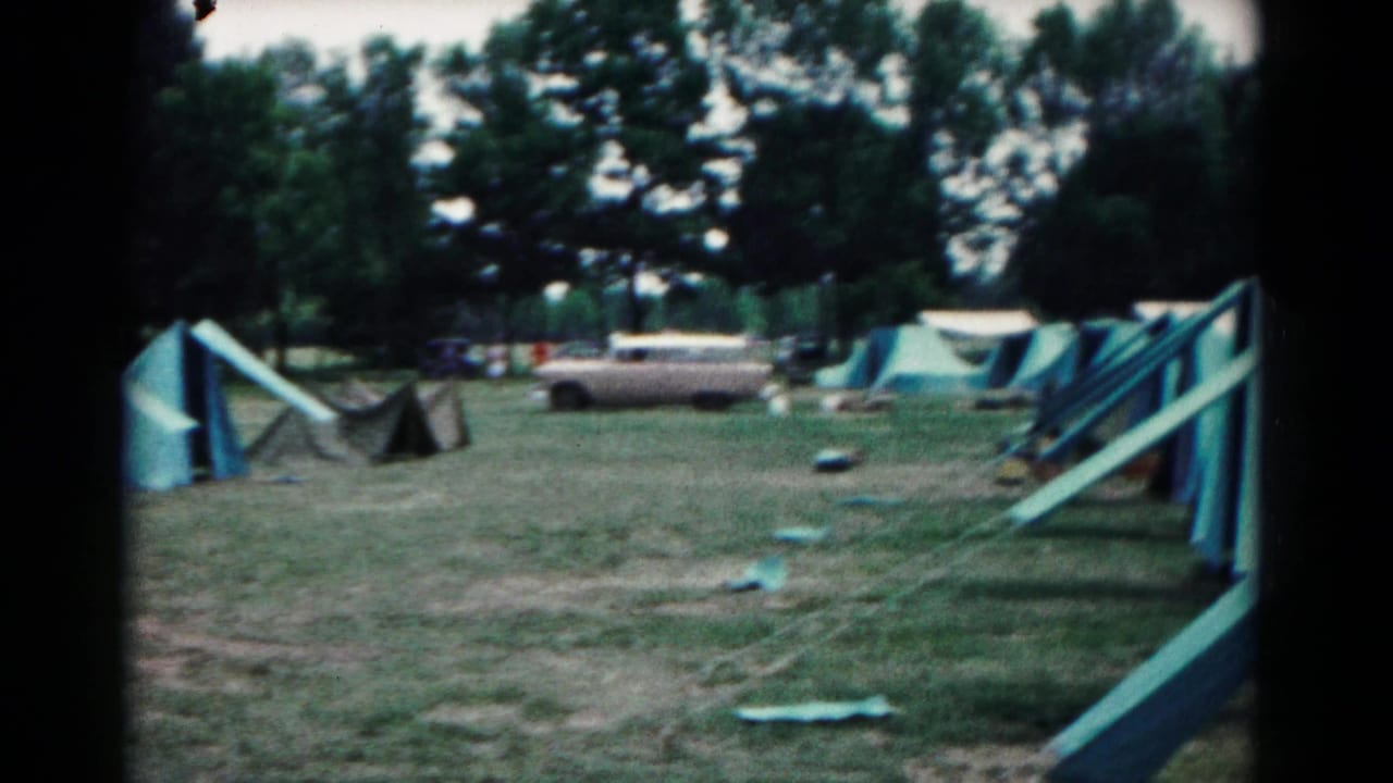 A 1966 home-movie frame shows tent rows at a Maryland campsite, with a parked station wagon, scattered gear, and dense trees beyond.
