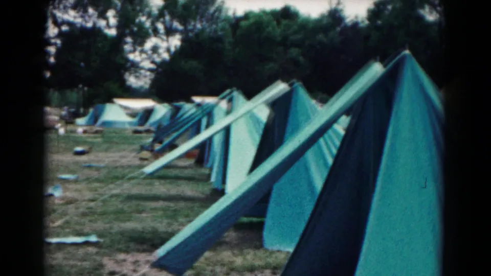 Close 8mm frame of blue-green canvas tents at the Hagerstown campsite, looking along guy lines and tent sides toward trees and distant camp gear.