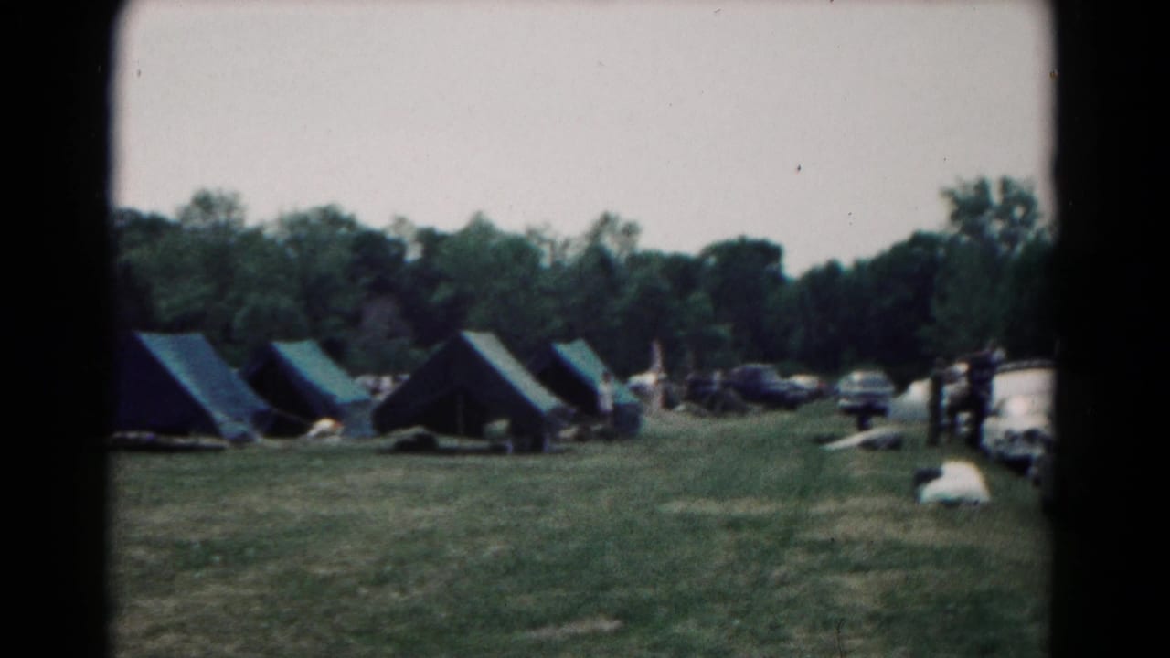 Grassy campsite with blue tents, parked cars, distant people, and a treeline.