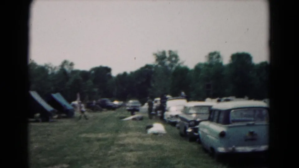 1966 Hagerstown camping scene with canvas tents, a line of 1960s cars, distant people, and trees behind the field.