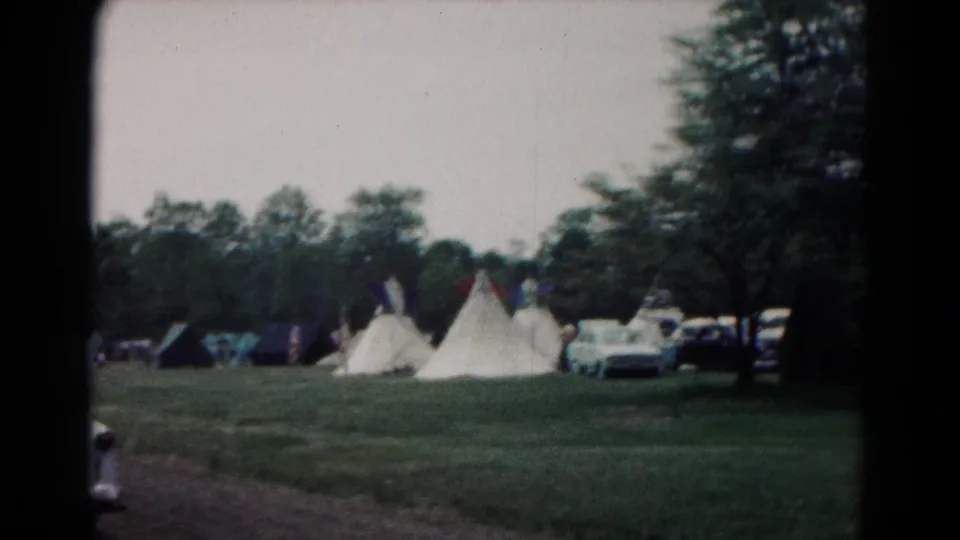1966 Hagerstown camping frame with white teepee-style tents, a parked car, darker tents, grass, and a wooded edge.