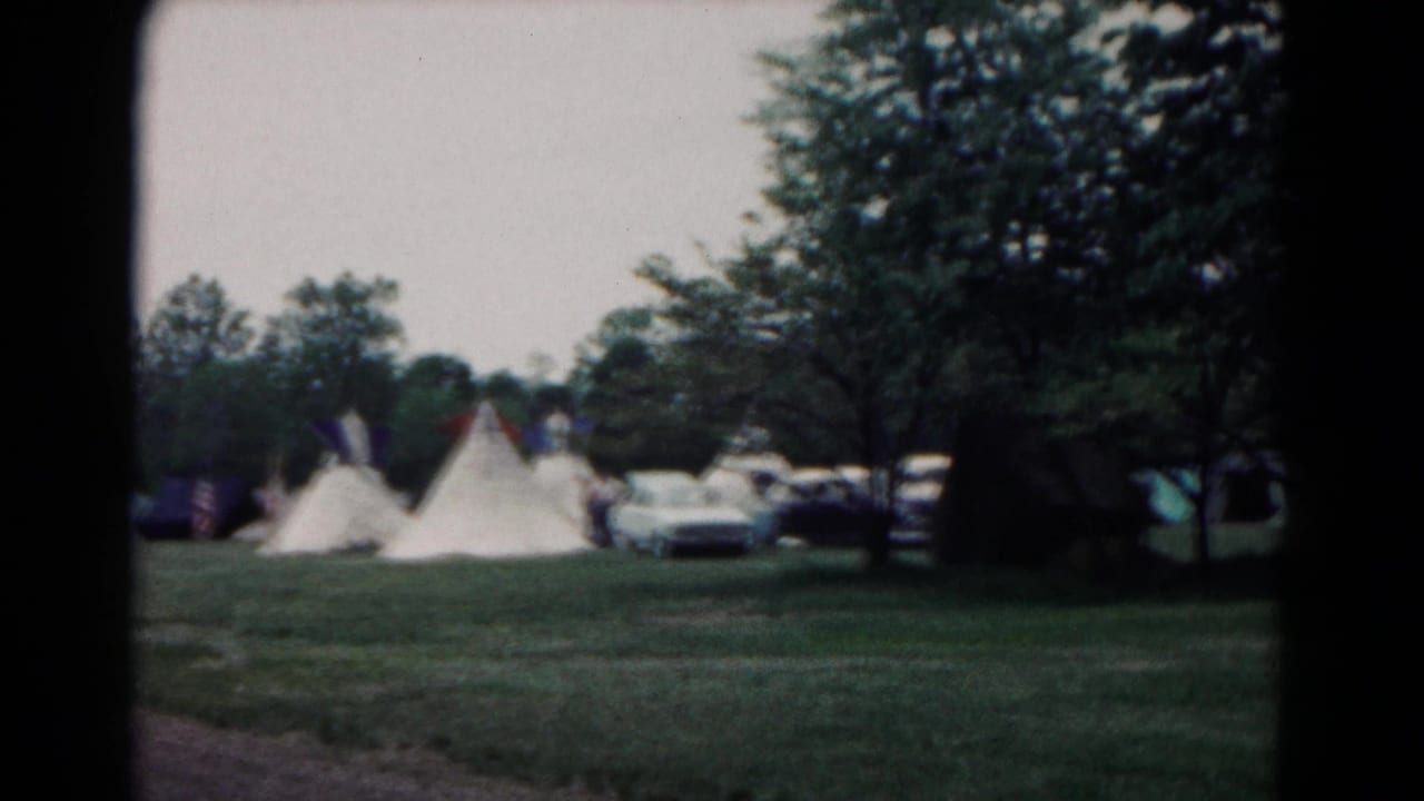Grassy campsite with white canvas tents, trees, and parked vintage cars in the background.