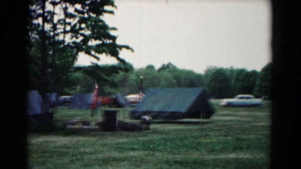1966 Hagerstown campsite frame with a dark canvas tent, nearby flags, parked cars, trees, and a person at frame edge.