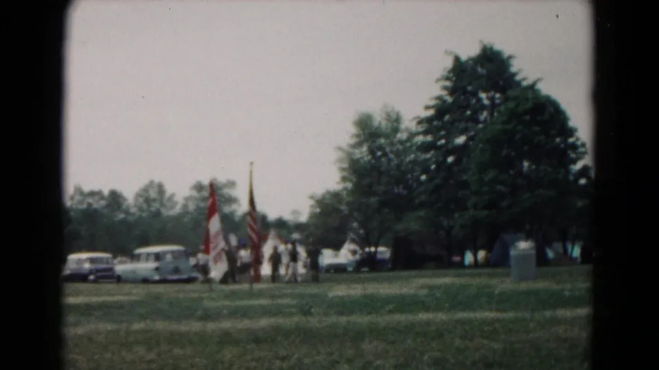 1966 Hagerstown home-movie frame showing distant people with flags near parked cars, trees, and open campsite grass.