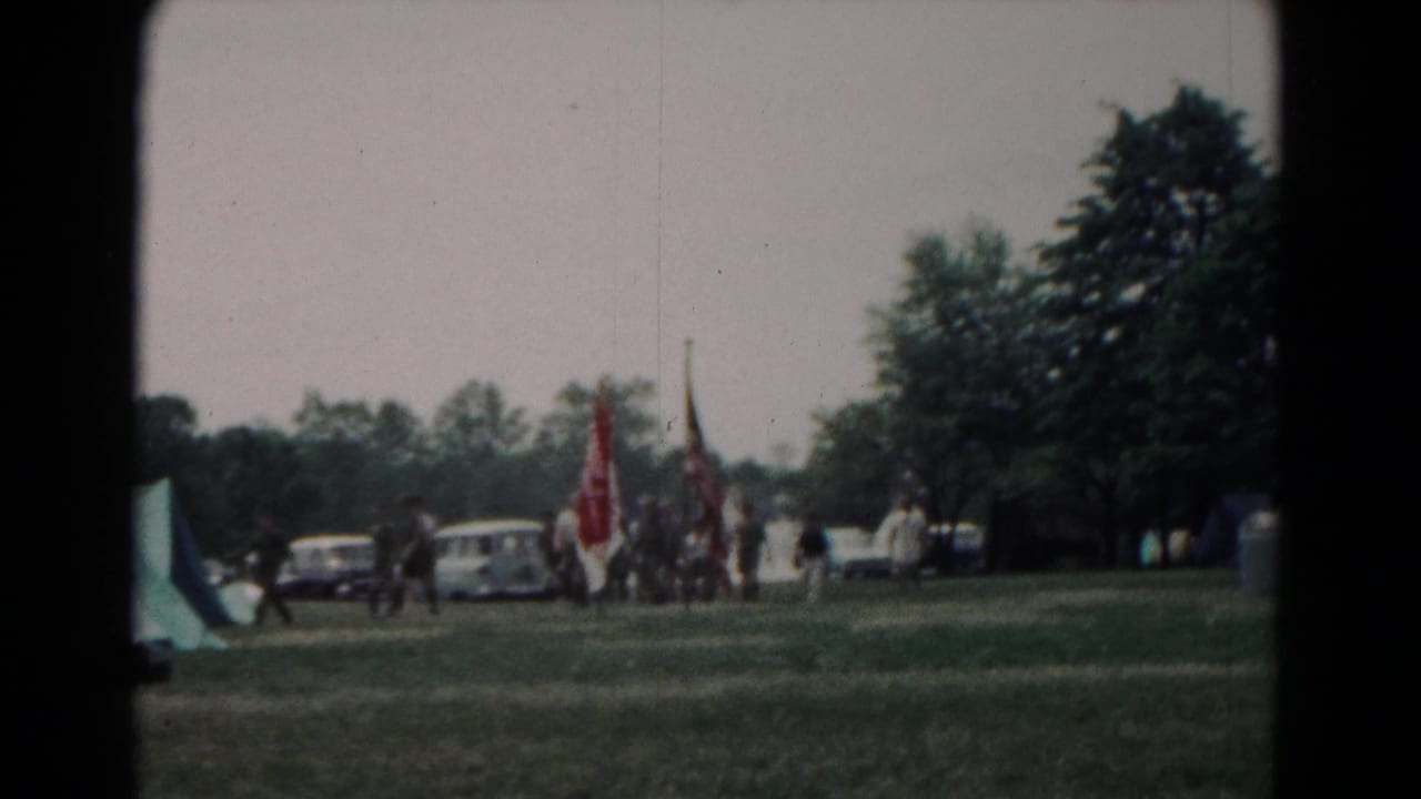 People walking with flags across a grassy field near tents and parked cars.