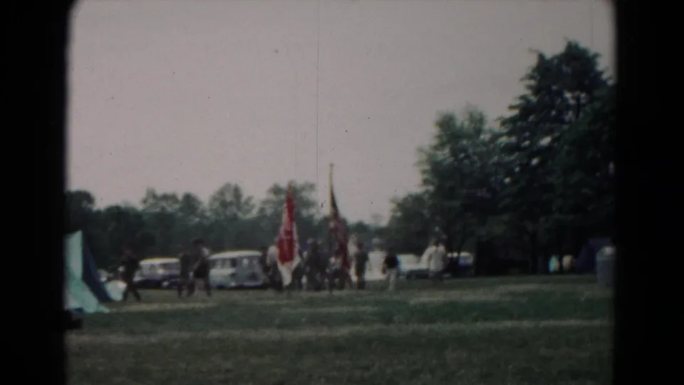 1966 Hagerstown home-movie frame showing people carrying flags across grass near tents, trees, and period cars.