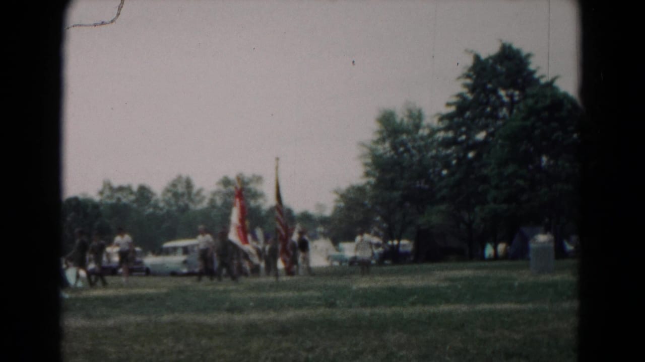 Flag-bearing group walks across a grassy Hagerstown field in 1966, with tents, parked cars, and trees in the background.
