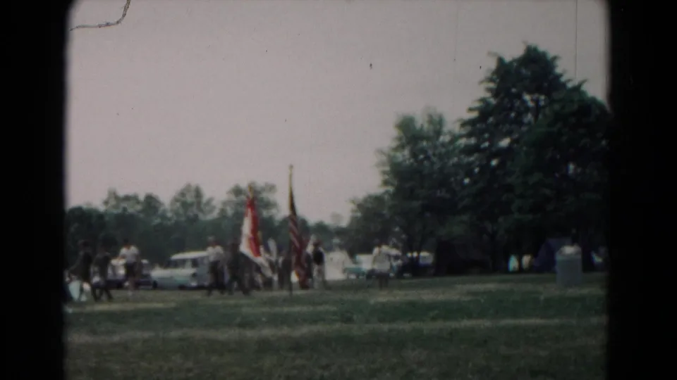 Flag-bearing group walks across a grassy Hagerstown field in 1966, with tents, parked cars, and trees in the background.