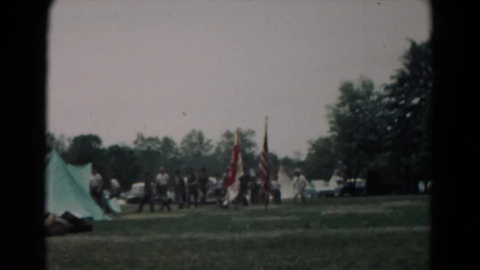 1966 Hagerstown outdoor-event frame with walkers and flags crossing grass near tents, trees, and distant cars.