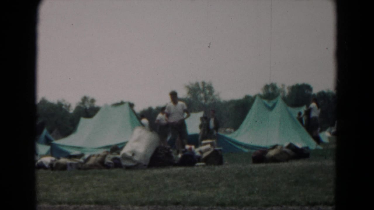 Green tents on a grassy campsite with campers and supplies nearby.